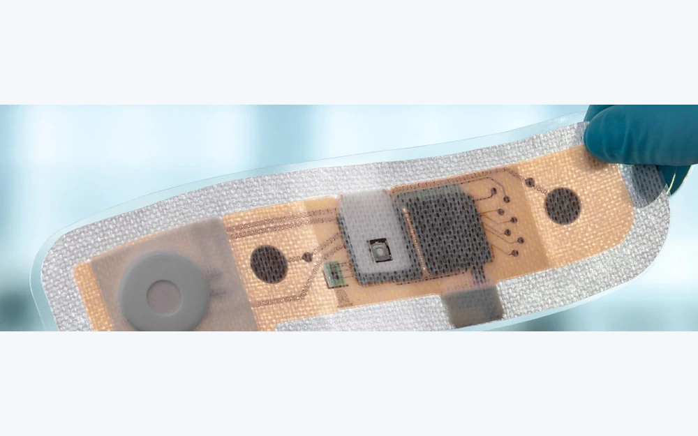 Parche sensor médico flexible con capas textiles, de circuito impreso y electrónicas visibles, sostenido por una mano enguantada