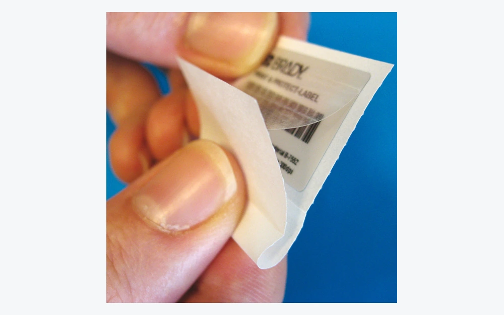 Hands lifting a clear protective film over a printed label on its release liner